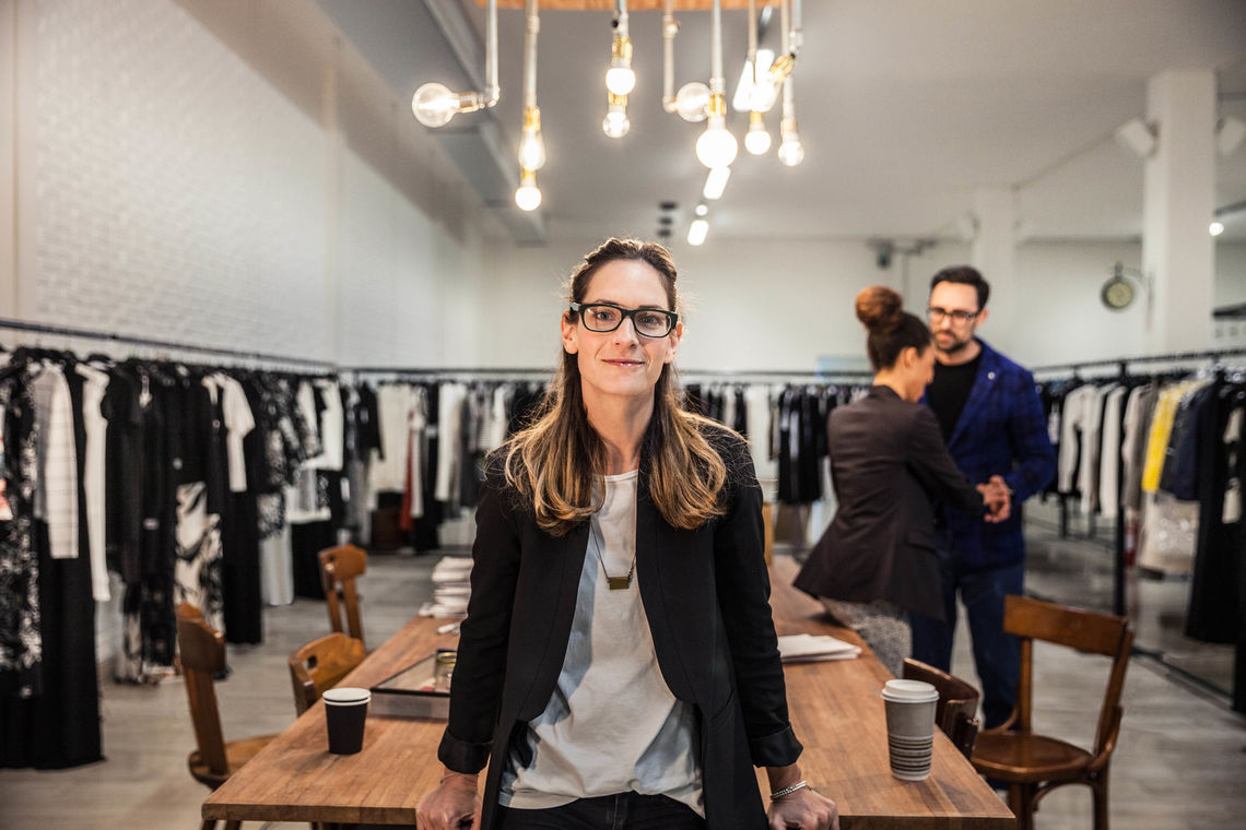 Eine im Business-Look gekleidete Frau steht in der Mitte des Bildes und sieht in die Kamera. Im Hintergrund sind verschiedene Kleiderstangen erkennbar.