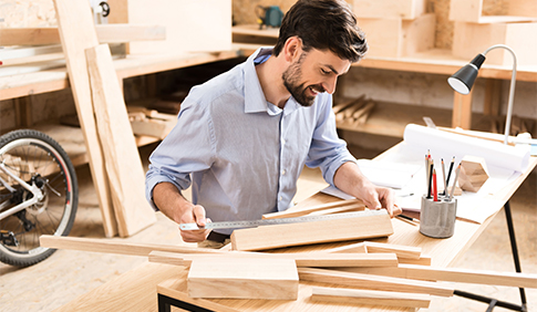 Ein Handwerker sitzt an einem Arbeitsplatz in einer Werkstatt. Er begutachtet ein Stück Holz. Im Hintergrund ist Holz zu sehen.